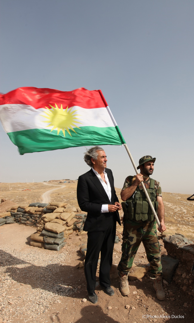 BHL dans le désert du Kurdistan, sur le front. Un militaire à côté de BHL porte un drapeau du Kurdistan irakien
