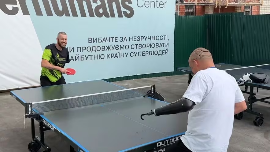 Les blessés de guerre dans un hôpital de Lviv. on voit un manchot jouer au ping pong avec un homme ayant une prothèse à la place du bras ainsi que de larges cicatrices sur l'arrière de son crâne.