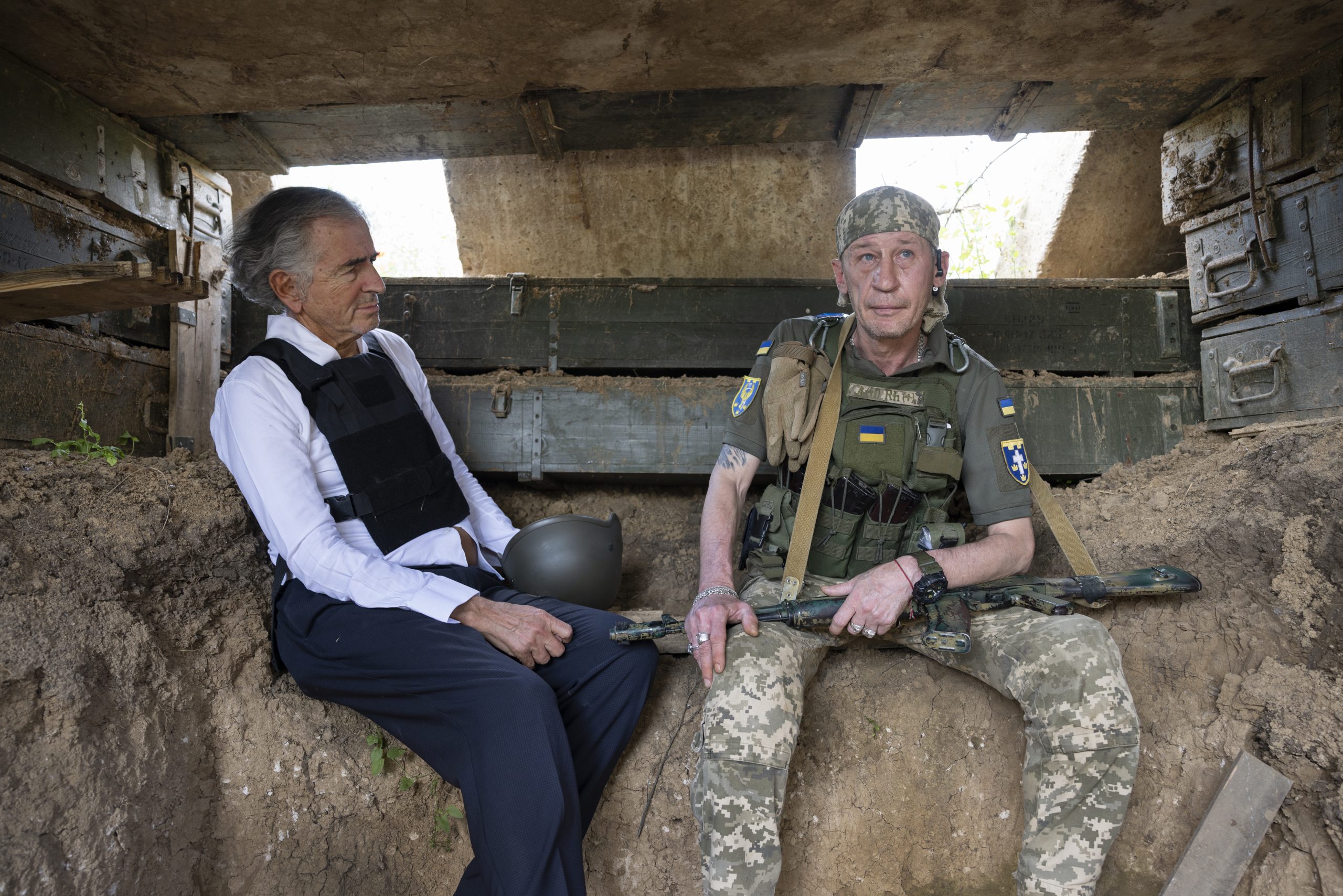 Bernard-Henri Lévy et un membre d'une unité d'assaut ukrainienne, au début de l'été 2023. Ils sont assis dans une tranchée.