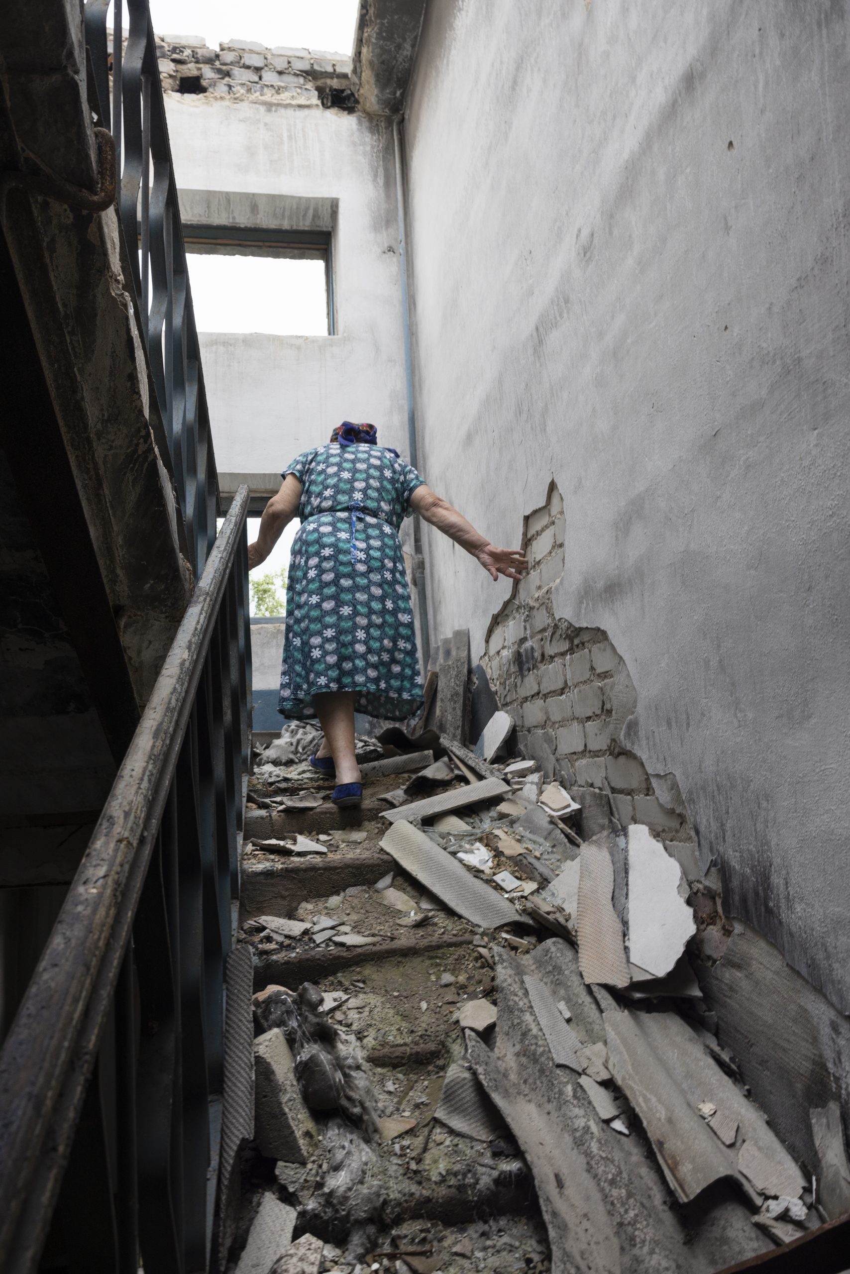 Un femme âgée, de dos, portant une robe à fleurs, monte l'escalier au milieu des débrits de la maison ravagée par un bombardement.