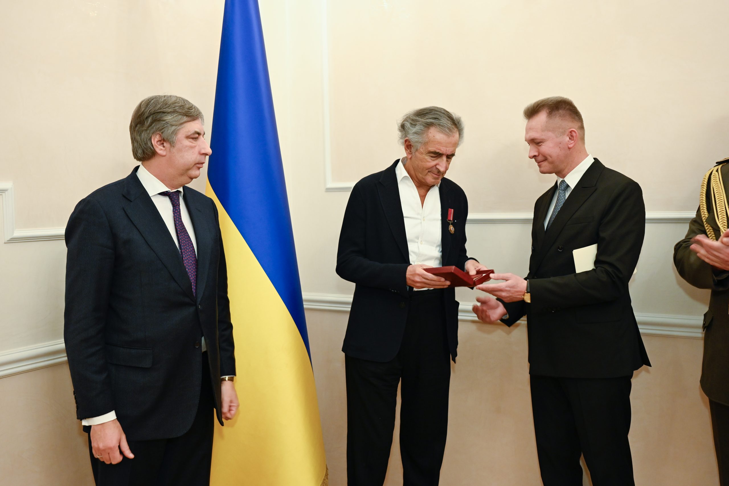 BHL reçoit l'insigne de Chevalier de l'Ordre du Mérite d'Ukraine. Il y a Vadym Omelchenko a côté de lui. Ils sont devant un drapeau ukranien.
