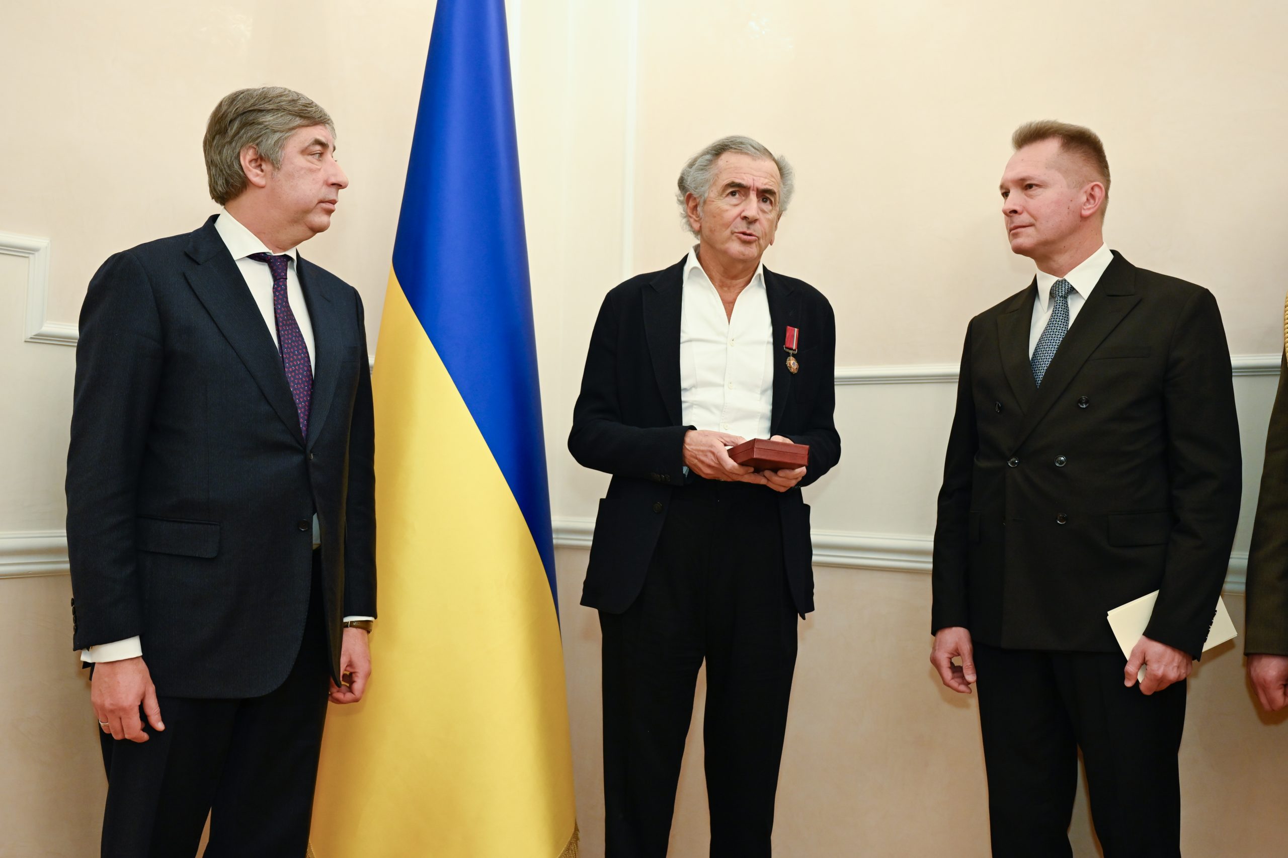 BHL reçoit l'insigne de Chevalier de l'Ordre du Mérite d'Ukraine. Il y a Vadym Omelchenko a côté de lui. Ils sont devant un drapeau ukranien.