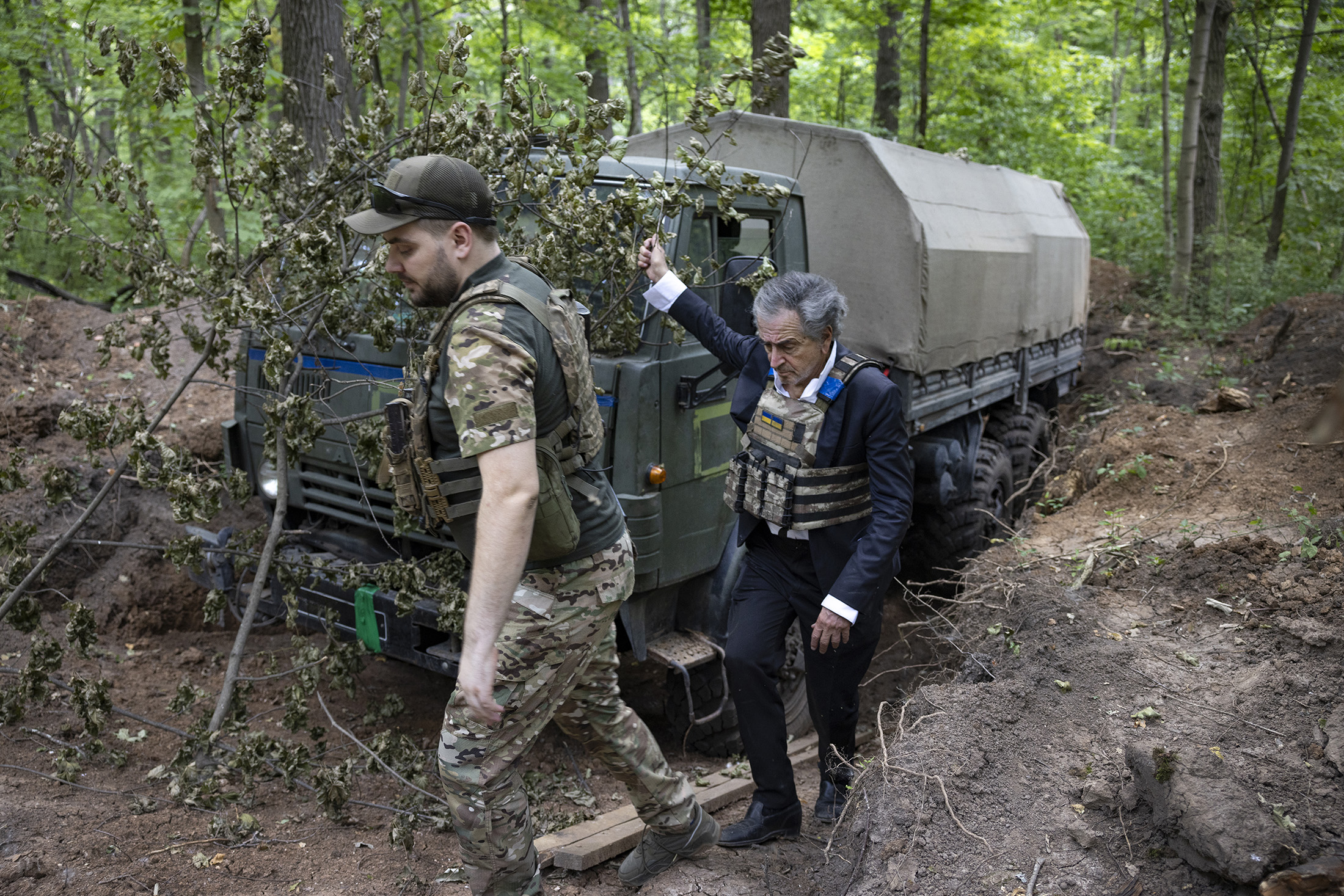 Bernard-Henri Lévy porte un gilet pare-balles, il est avec un militaire ukrainien dans une forêt près d'un camion de l'armée.