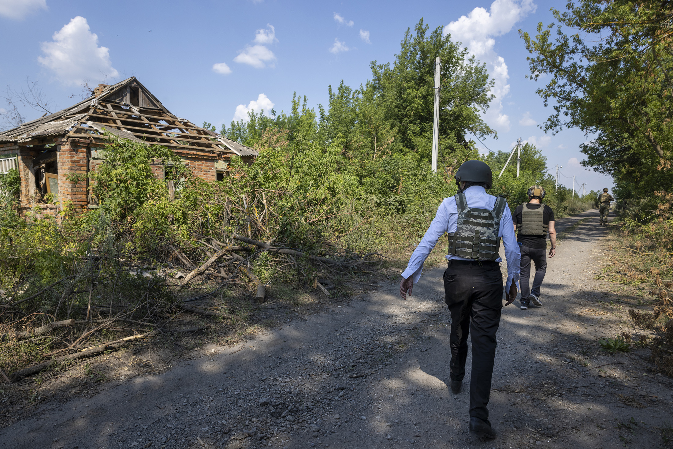 BHL de dos, il porte un gilet pare-balles et un casque militaire, il marche sur un chemin de campagne entouré de maisons en ruines