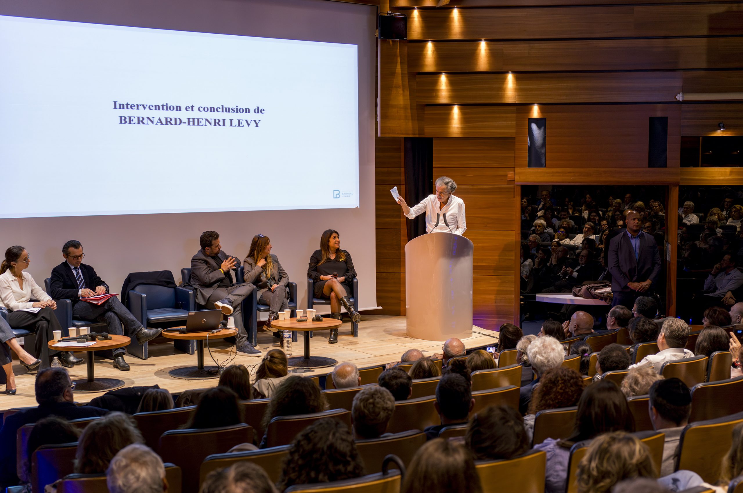 BHL parle derrière un pupitre sur la scène de l'auditorium de la Maison du Barreau, devant les co-Présidentes de la Commission ouverte Paris Israël du Barreau de Paris, et un collectif d'avocats.