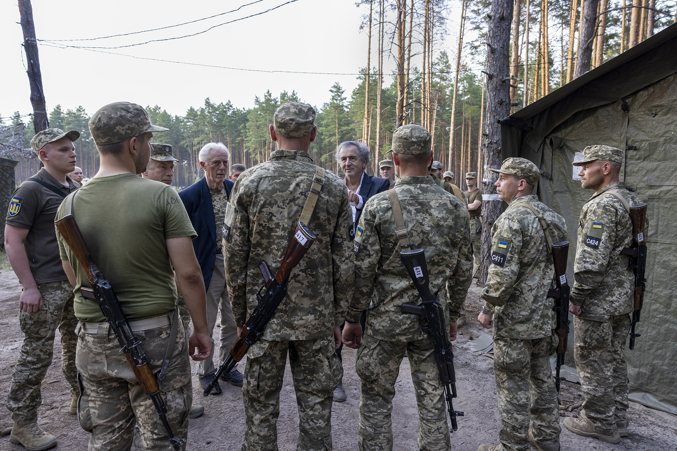 BHL et Gilles Hertzog dans un camp militaire, dans une forêt au nord de Kharkiv. Ils parlent avec des soldats en uniforme.