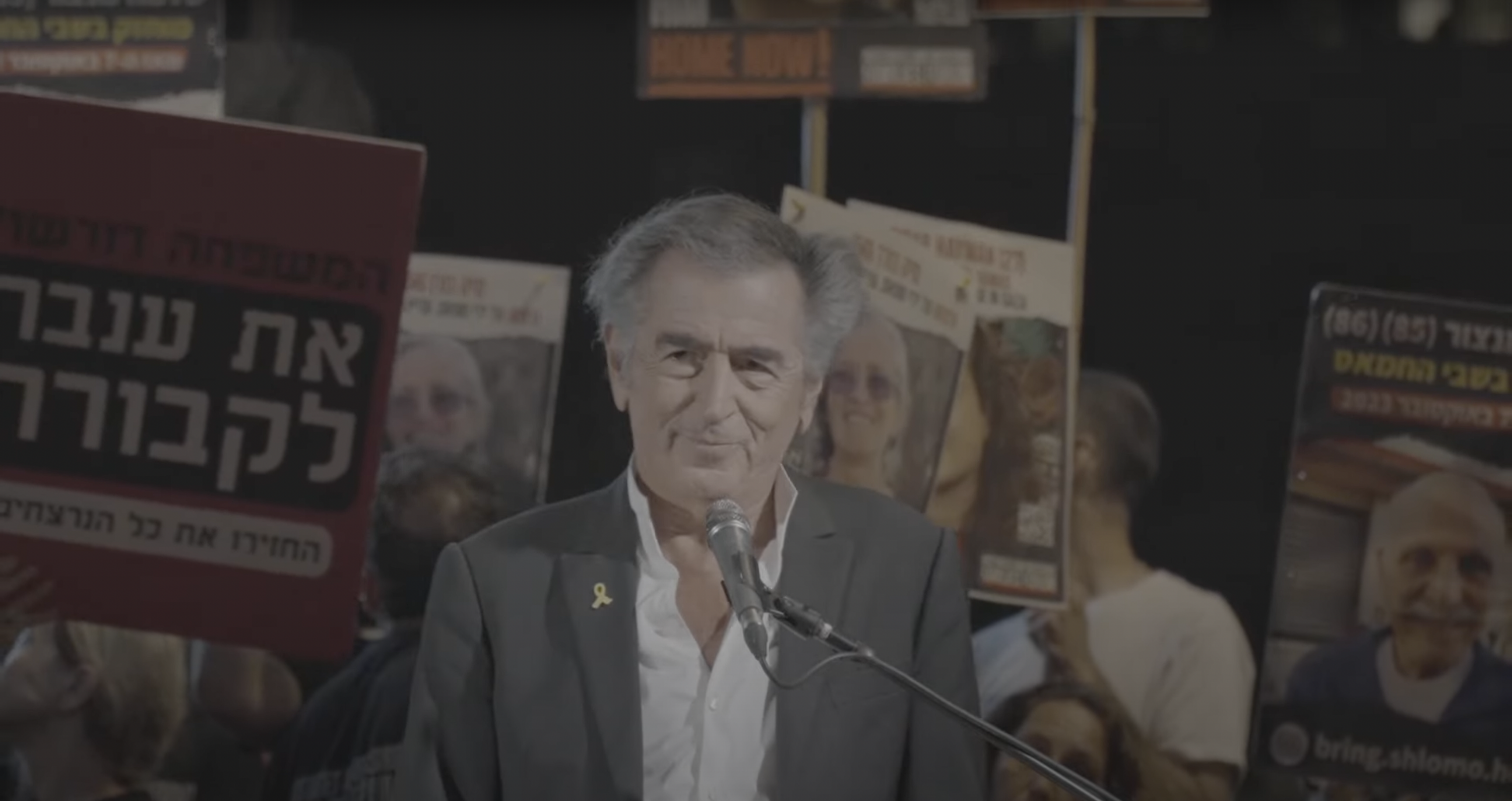 Bernard-Henri Lévy fait un discours sur la Place des Otages à Tel-Aviv, en Israël, le 11 mai 2024. Derrière lui on voit des hommes et des femmes qui portent des pancartes avec des visages d'otages israéliens.