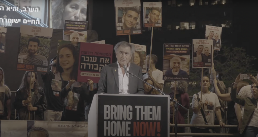 Bernard-Henri Lévy fait un discours sur la Place des Otages à Tel-Aviv, en Israël, le 11 mai 2024. Derrière lui on voit des hommes et des femmes qui portent des pancartes avec des visages d'otages israéliens.