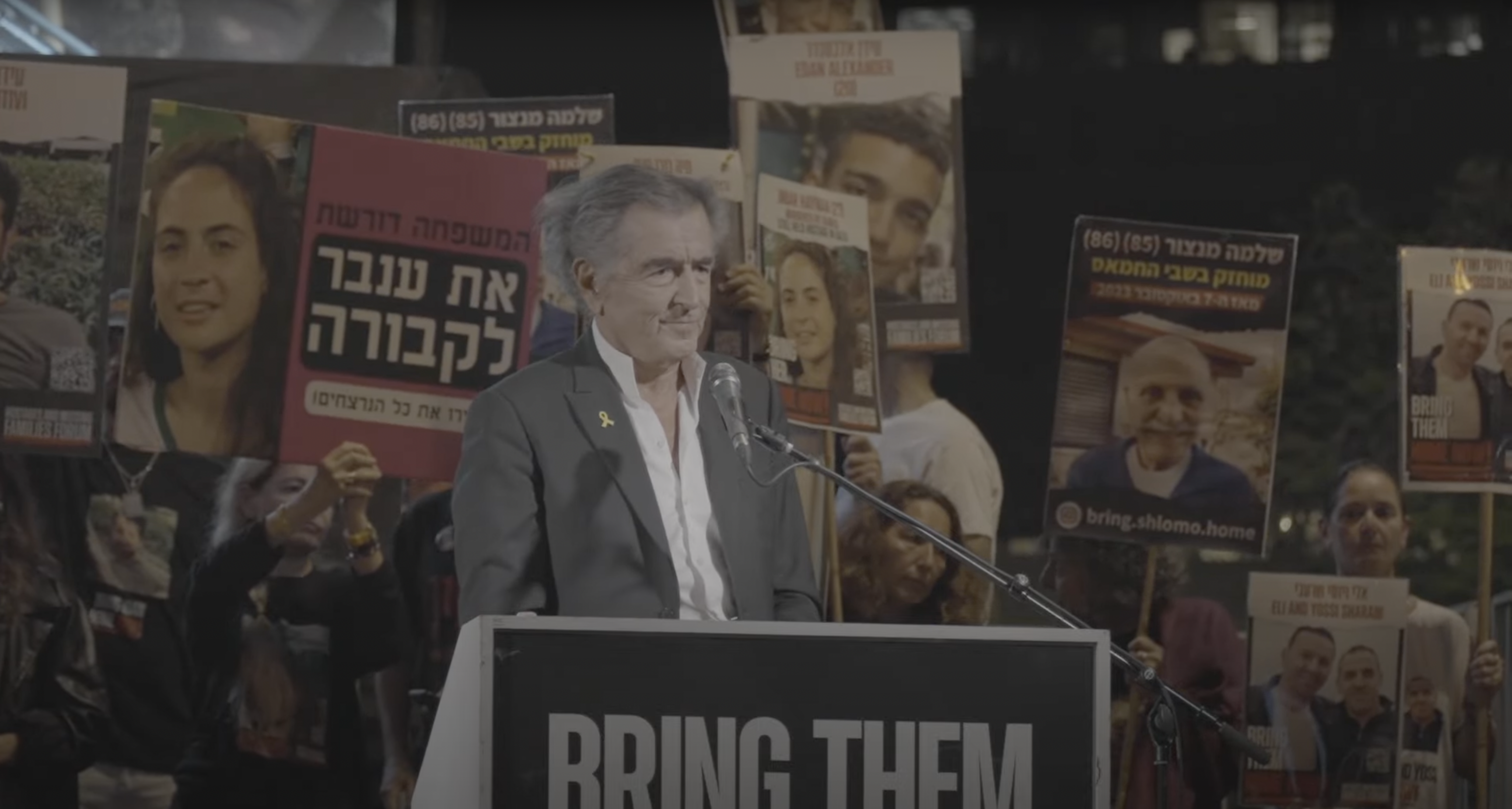 Bernard-Henri Lévy fait un discours sur la Place des Otages à Tel-Aviv, en Israël, le 11 mai 2024. Derrière lui on voit des hommes et des femmes qui portent des pancartes avec des visages d'otages israéliens.